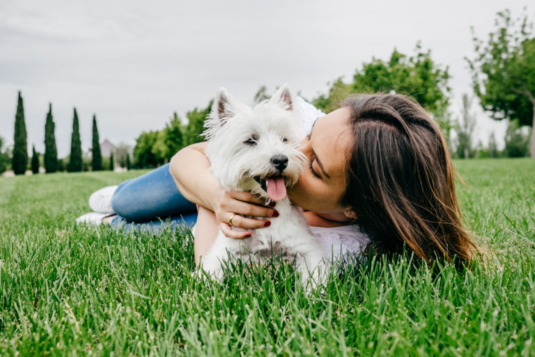 A woman laying on the grass kissing her small white cairn terrier